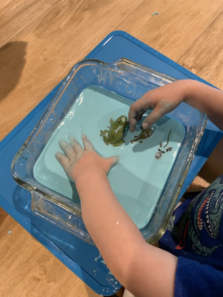 toddler hands playing with frog life cycle sensory bin with blue oobleck