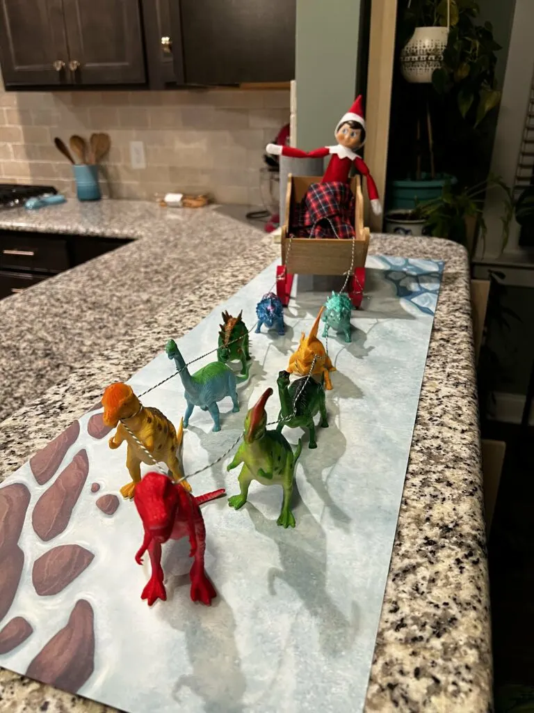 elf on the shelf in a sleigh with dinosaurs pulling it