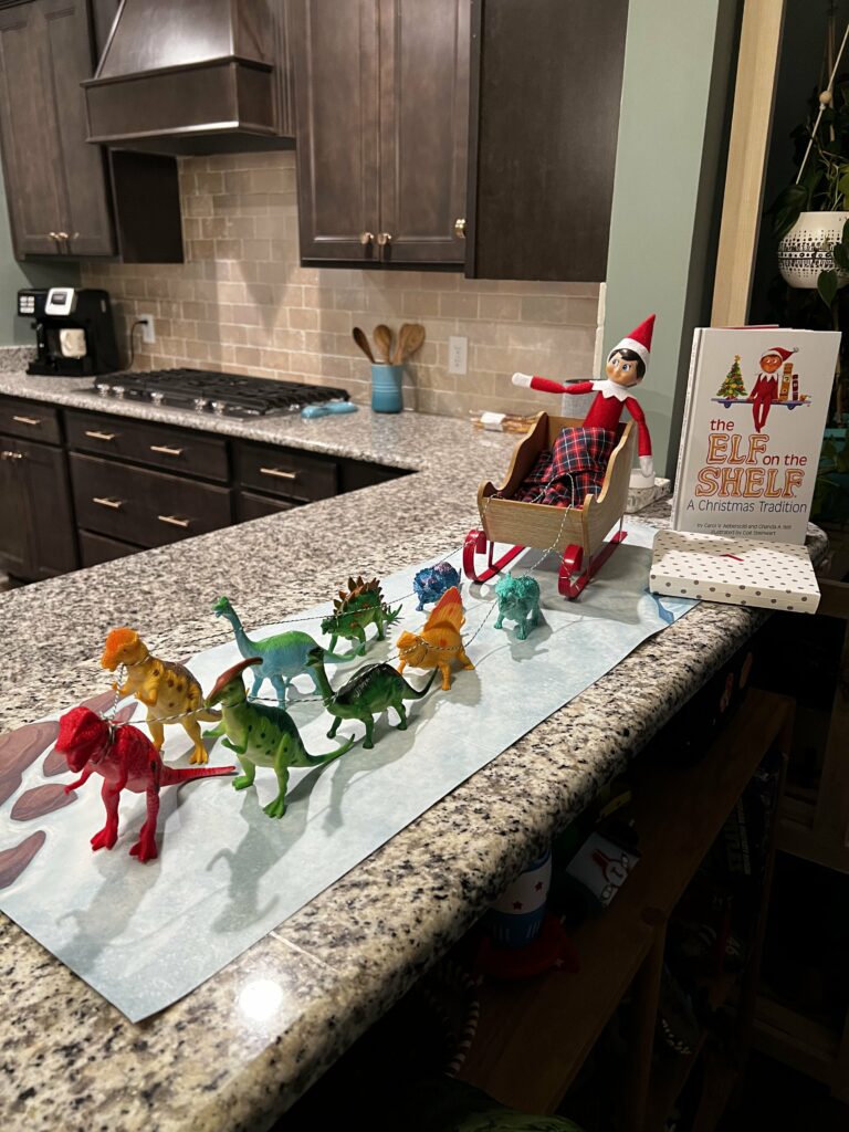 elf on the shelf in a sleigh with dinosaurs pulling it